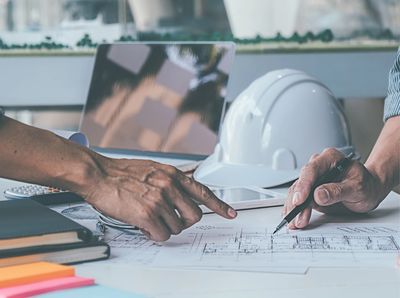 Hand pointing to architectural plans on a table with a hard hat and laptop.