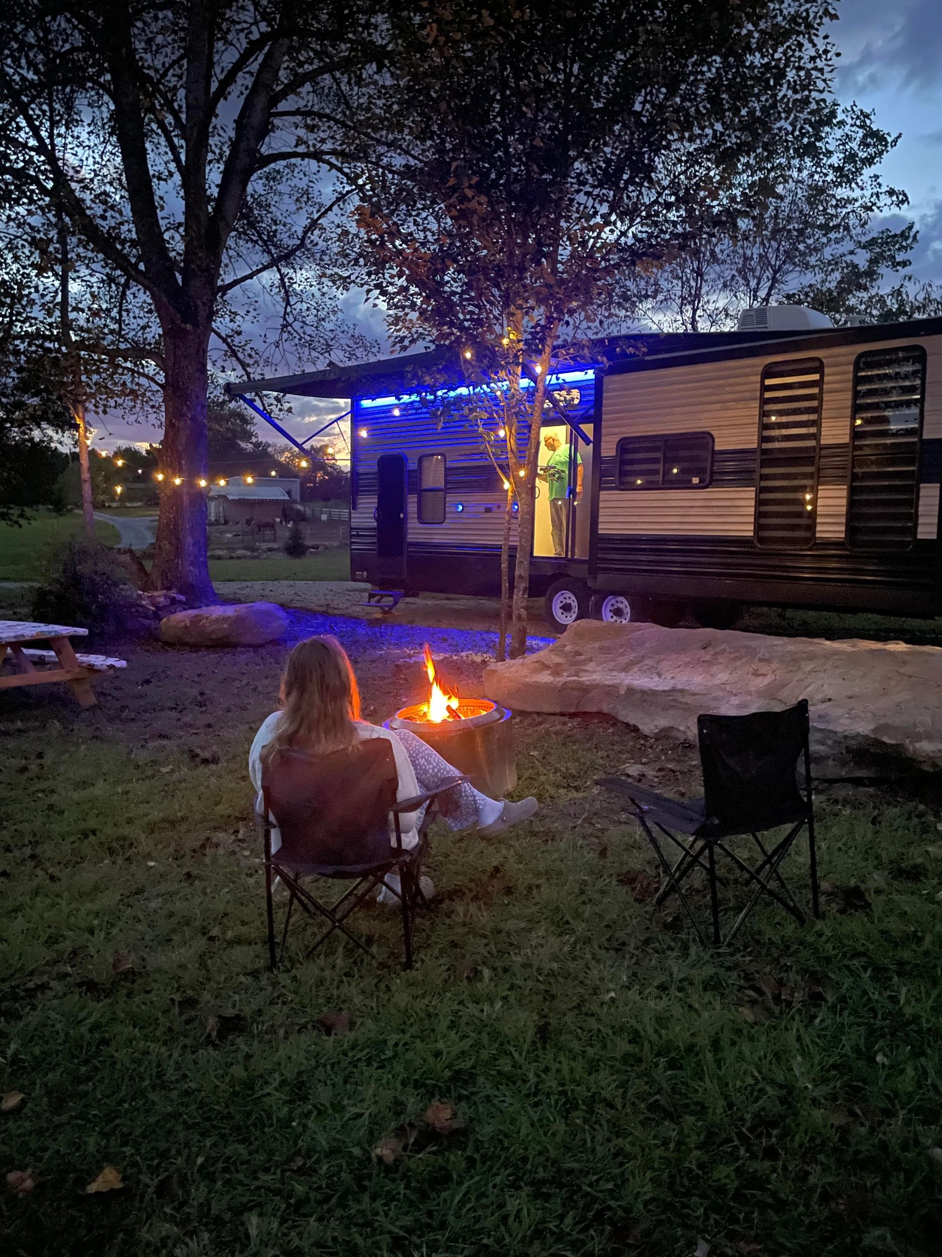 Person relaxing by a campfire near a lit RV at dusk.