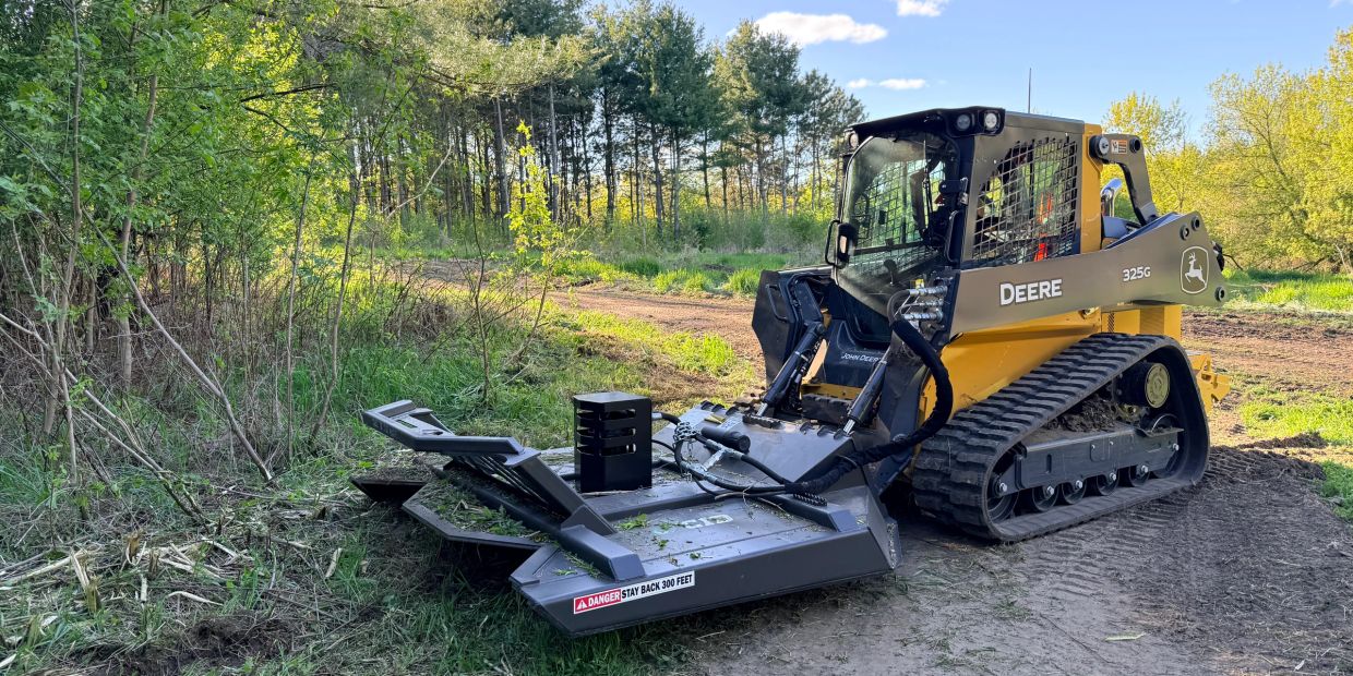 John Deere 325G tracked skid steer with forestry mulcher in a wooded area.