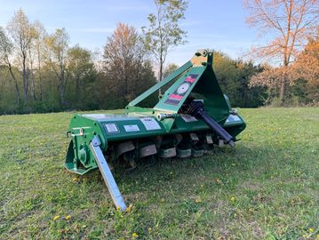 Green agricultural tiller machine on grassy field with trees in background.