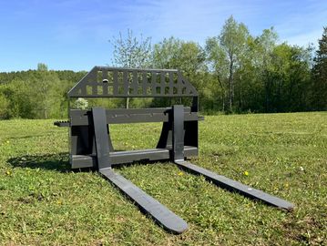 Metal forklift attachment on grass with trees and blue sky.
