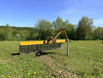 Yellow farming rake on green field with trees and blue sky.