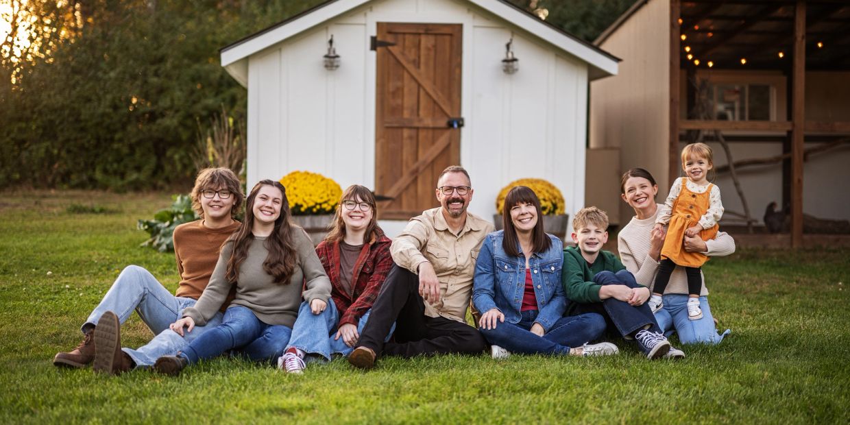 A happy family of eight sitting on grass in front of a white shed during sunset.