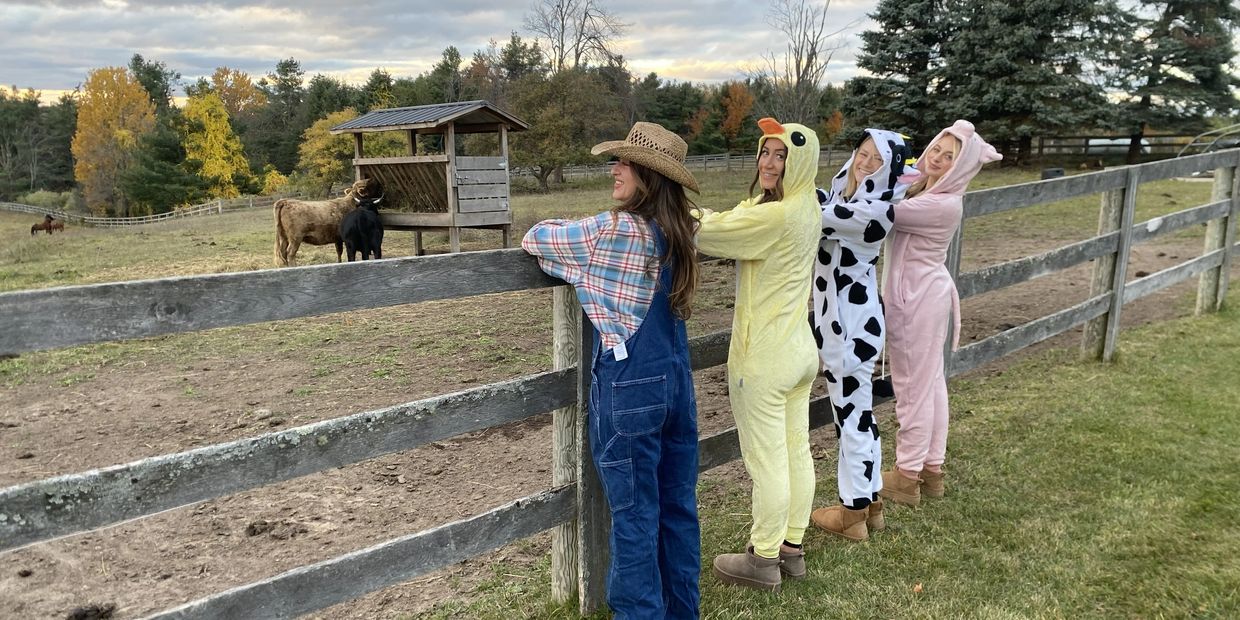 Four women in animal and farmer costumes stand by a wooden fence on a farm.