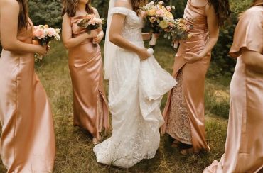 Bride in white gown with bridesmaids in satin pink dresses holding flowers outdoors.