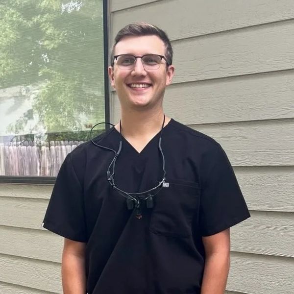 Smiling man in black scrubs with glasses and dental loupe headset.