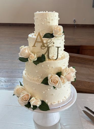 Elegant four-tier white wedding cake adorned with cream roses and gold initials.