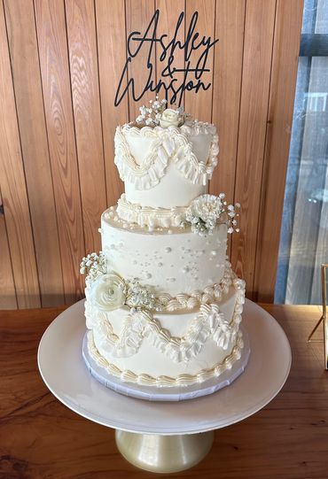 Elegant three-tier white wedding cake with floral decorations and custom name topper.
