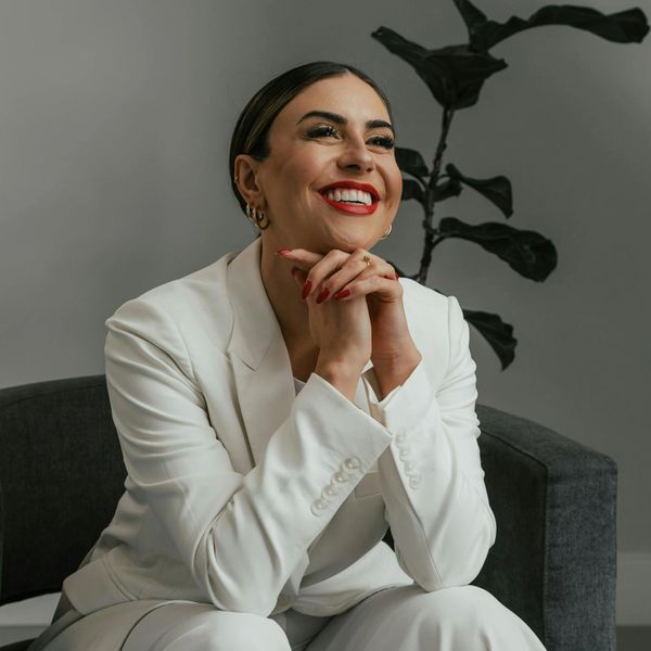 Confident woman in a white suit smiling brightly, sitting on a dark chair.