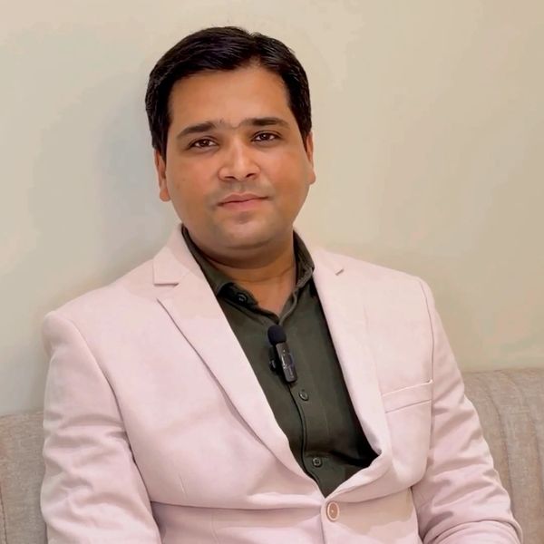 A man in a light-colored blazer and dark shirt sitting and smiling.