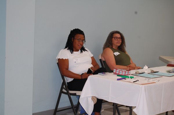 Two women sitting at a table with papers and clipboards.