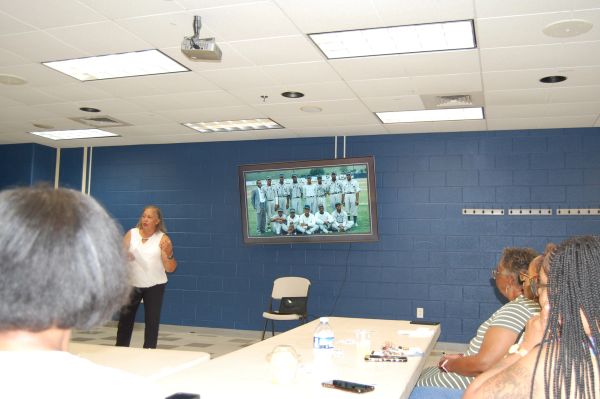 Woman speaking to group in meeting room with sports photo on TV.