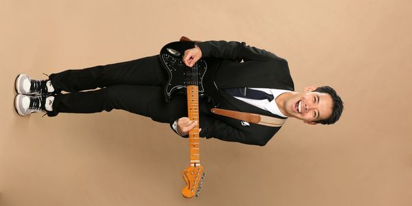 A man in a suit joyfully playing an electric guitar against a beige background.
