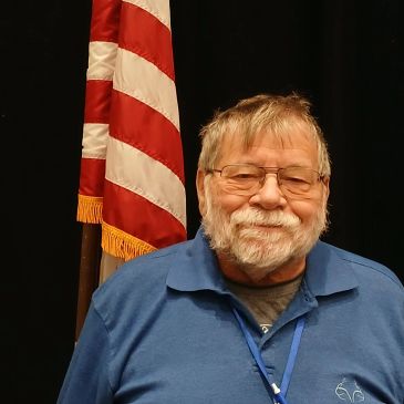 Elderly man with glasses smiling in front of an American flag.