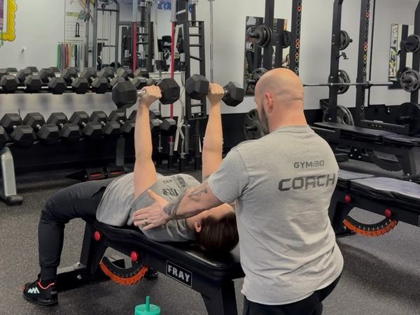 A coach assists a person lifting dumbbells on a bench in a gym.