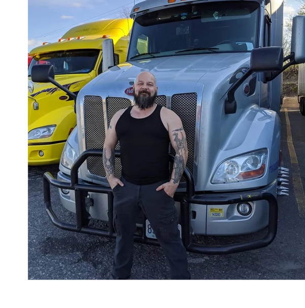 Bearded man with tattoos standing confidently in front of a silver Peterbilt truck.