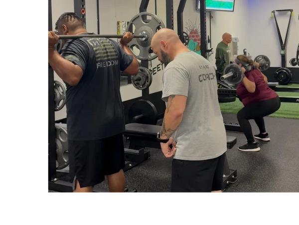 Two people lifting barbells while a coach supervises their squats in a gym.