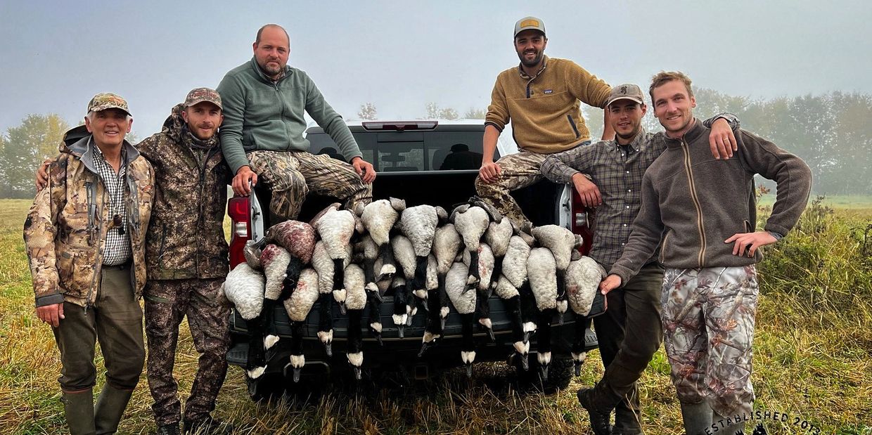 Six hunters pose with their goose hunt haul on a truck bed in a foggy field.