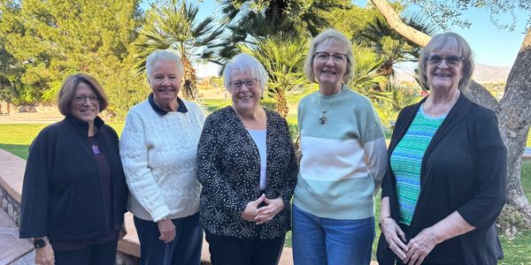 Five elderly women standing outdoors, smiling at the camera on a sunny day.
