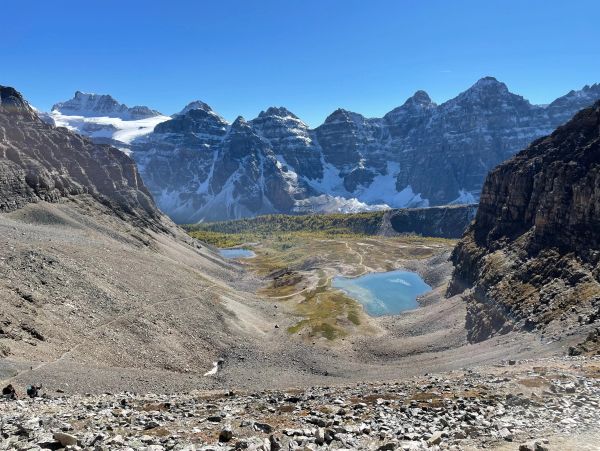 Rocky valley with small lakes beneath snow-capped mountains under a clear blue sky.