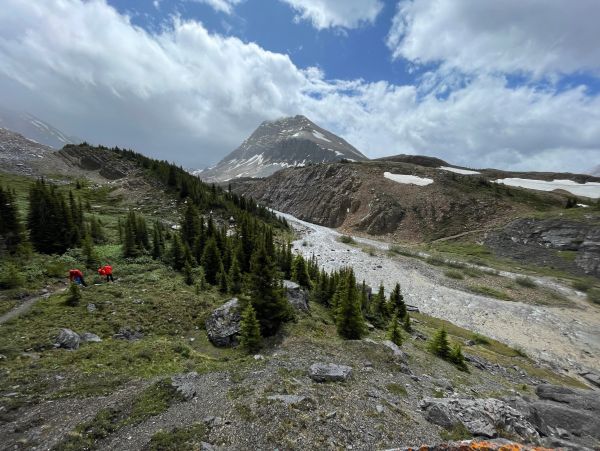 Two hikers explore a green alpine valley with a snow-capped mountain under a partly cloudy sky.