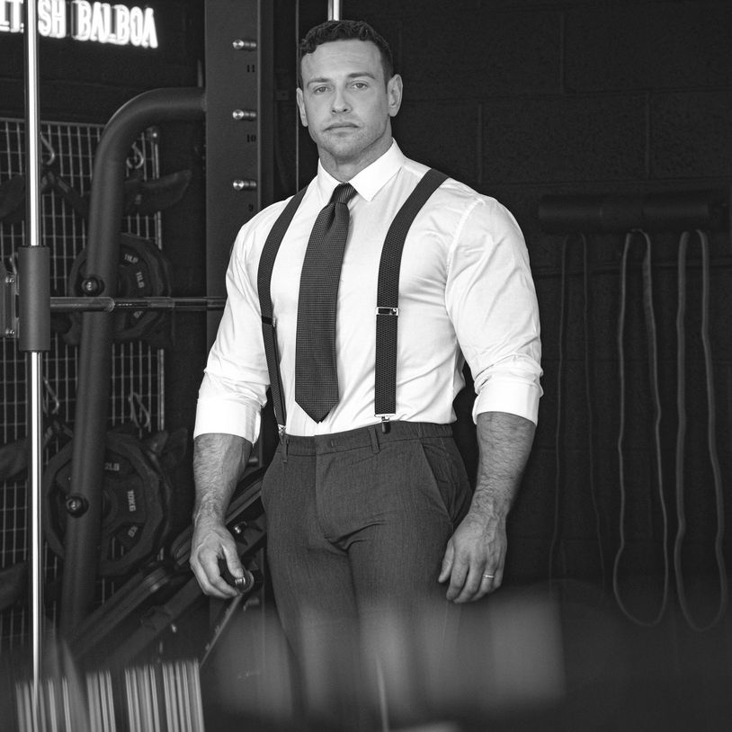 Muscular man in formal attire with suspenders in a gym setting.