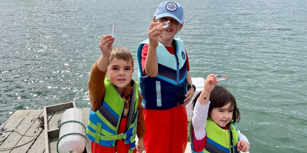 3 kids holding up vials of saltwater on a floating dock as part of an aquaculture field trip