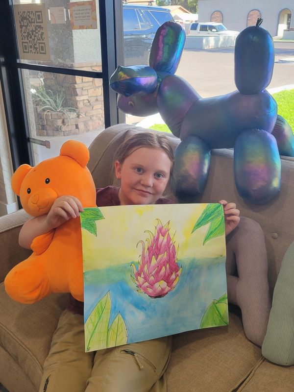 Child holding orange teddy bear and colorful drawing while sitting on couch with balloon dog.