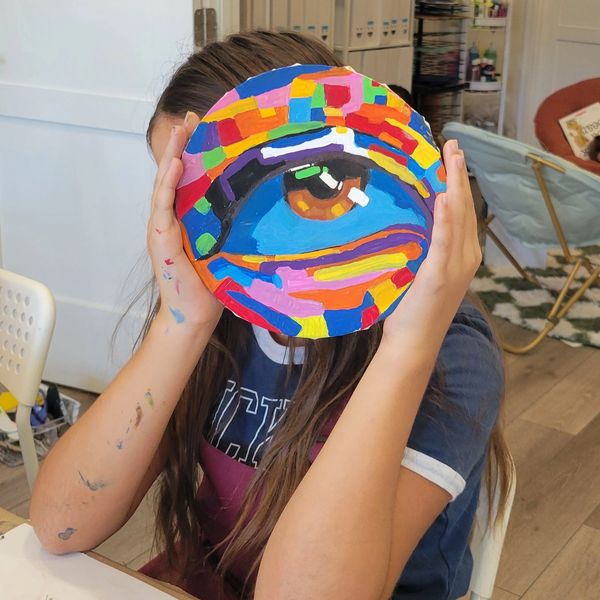 A person holds a colorful painted circle with an eye design in front of their face.
