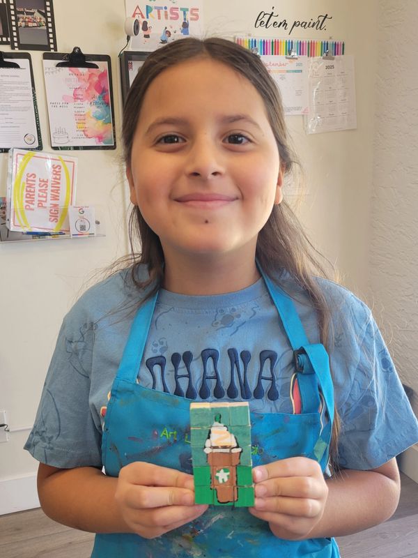 Smiling girl in paint-stained apron holding a painted wooden puzzle of a coffee cup.