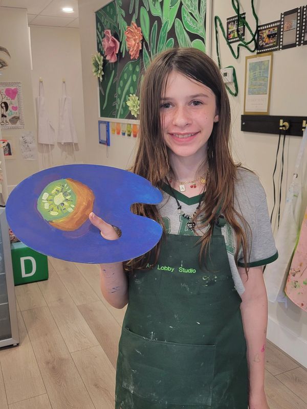 Young girl proudly displays a painted art palette in a creative studio.