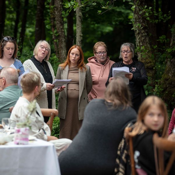 Group of women singing outdoors to an audience in a forest setting.
