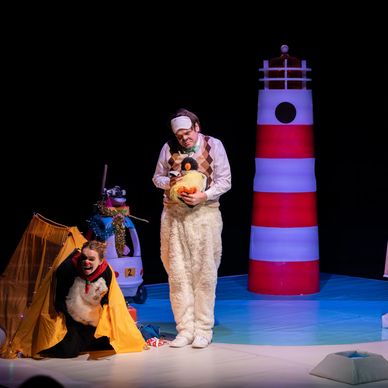 Two actors in animal costumes performing on stage near a lighthouse prop.