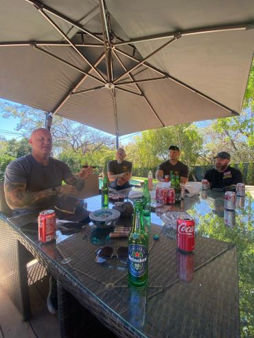 Four men sitting around a glass table outdoors under a large umbrella with drinks.