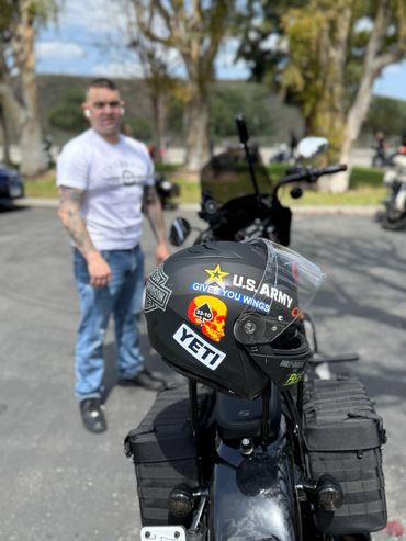 A motorcycle helmet with various stickers is placed on a bike, with a man standing blurred in the background.