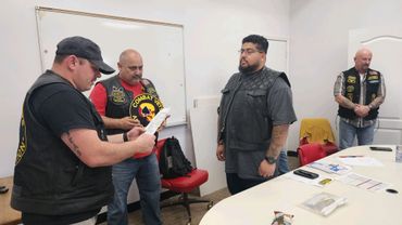 Four men in a room, two reading papers, wearing motorcycle club vests.