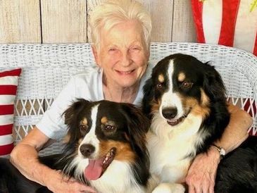 Elderly woman joyfully posing with two affectionate dogs on a white wicker bench.