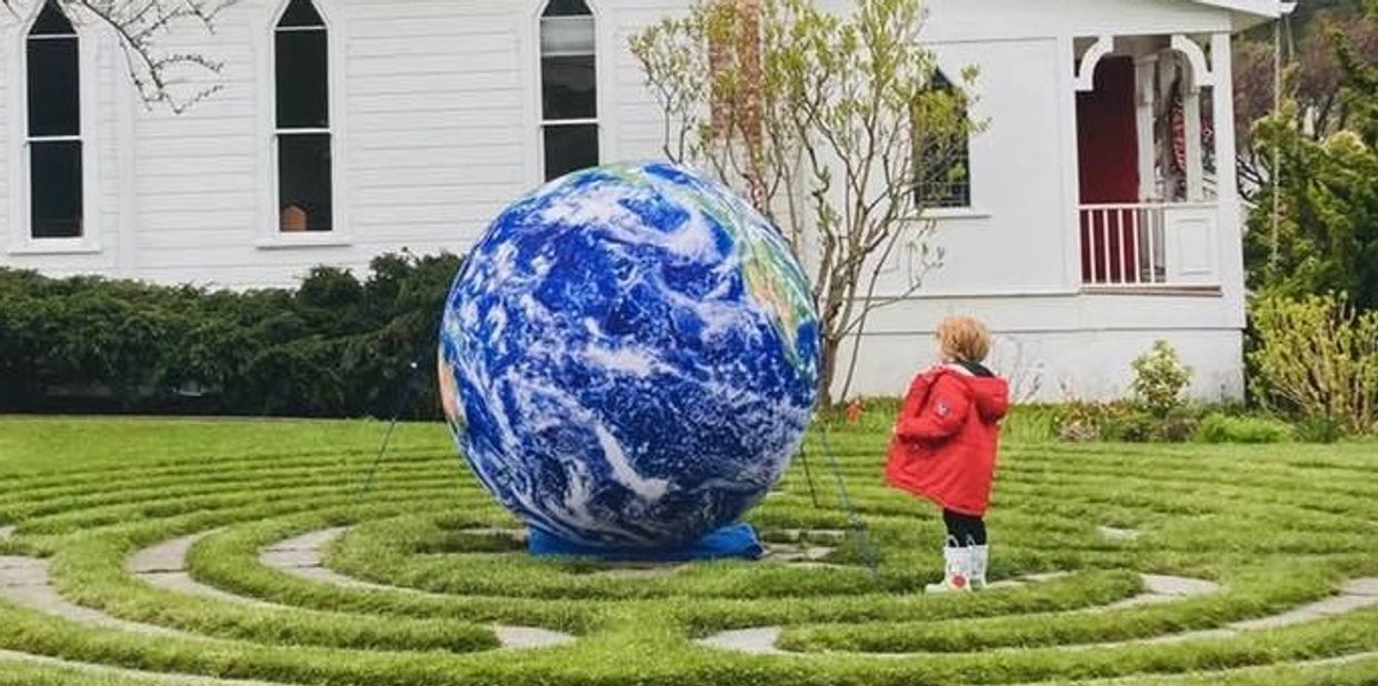 A child in a red coat stands near a large globe in a garden maze.