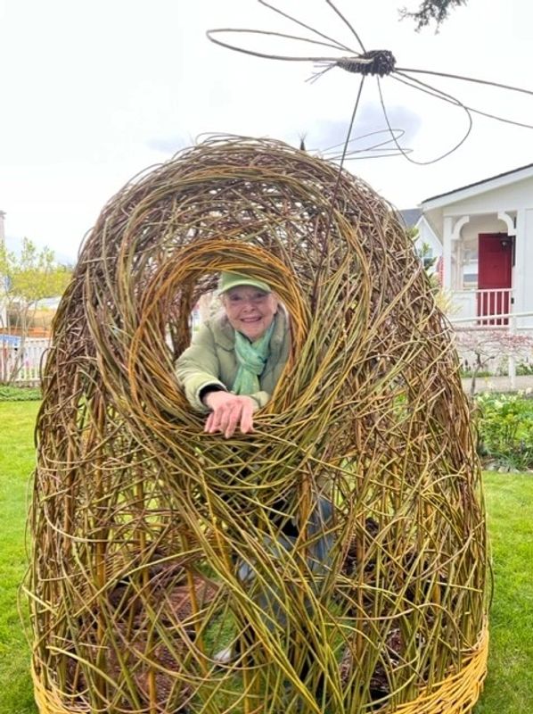 An elderly woman smiles inside a large woven twig sculpture with a dragonfly on top.