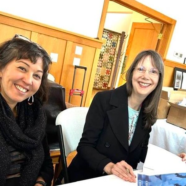 Two women smiling at a book signing event indoors.