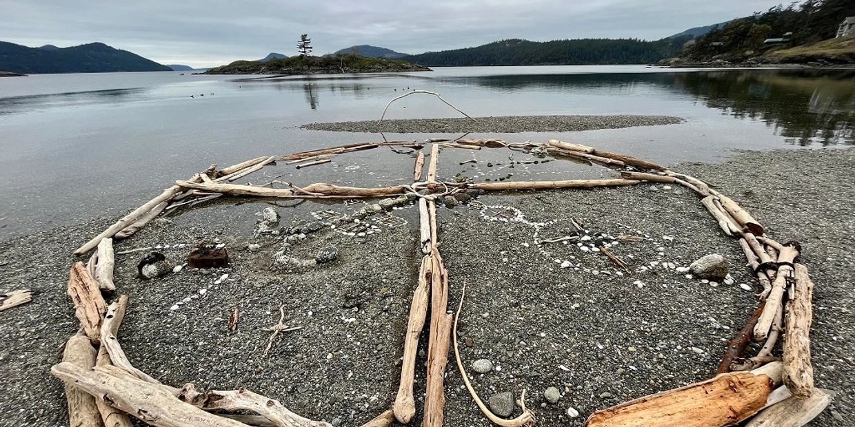 Driftwood and stones arranged in a circular pattern on a rocky shore with a calm lake and hills in the background.