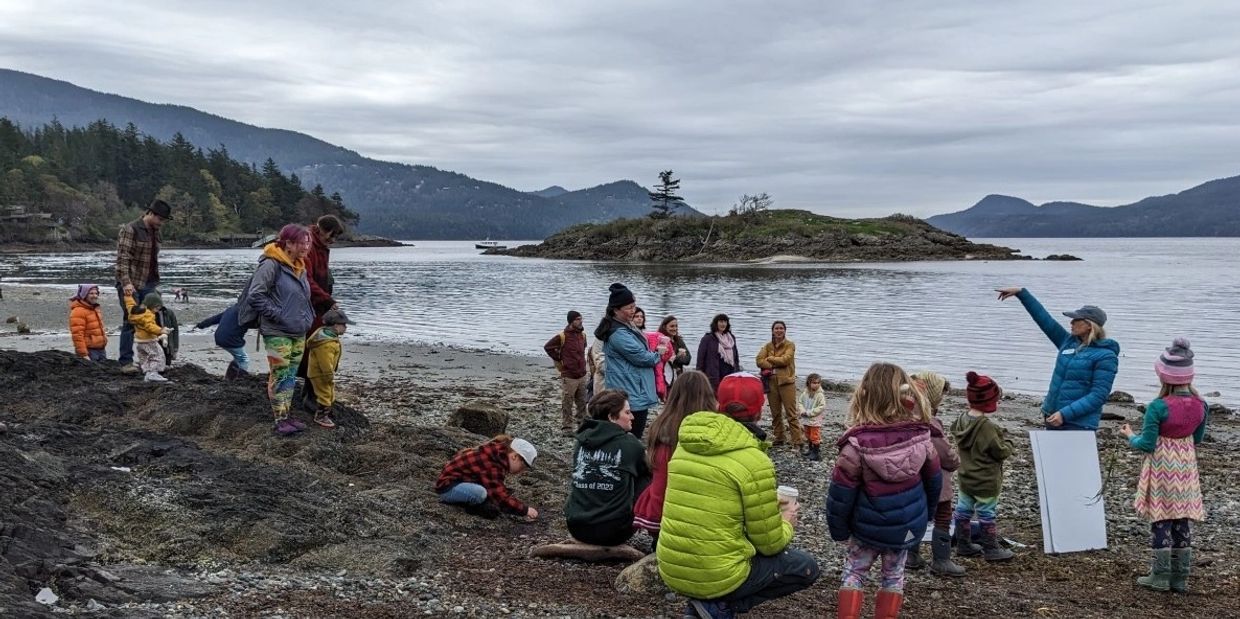 Group of people and children gathered on a rocky beach near a body of water and mountains.