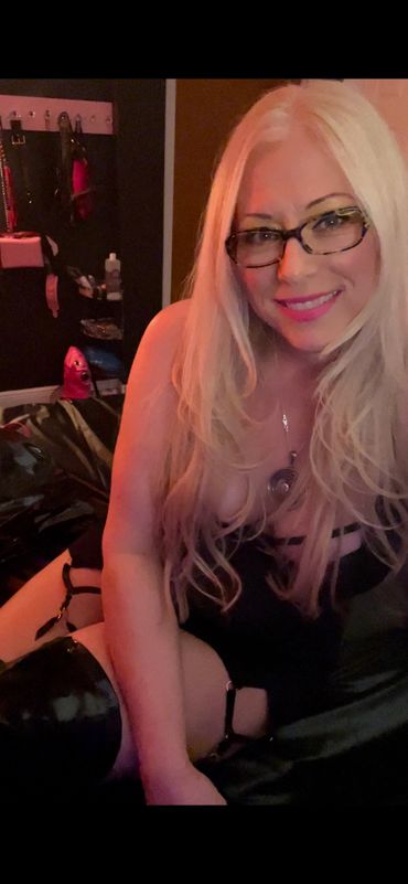Smiling woman with long blonde hair and glasses in a dimly lit room.