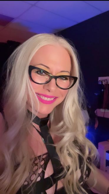 Smiling woman with long blonde hair and black glasses in a dark room.