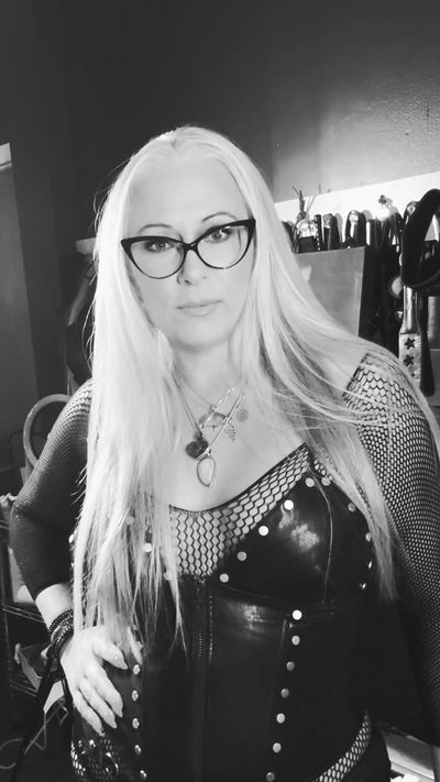 Woman with long blonde hair wearing glasses and a leather outfit in a dimly lit room.