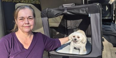 Woman sitting beside a small white dog in a portable pet crate.
