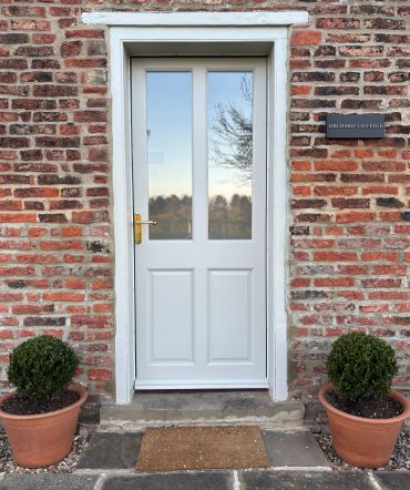 White door with glass panels on a brick wall, flanked by two potted bushes.