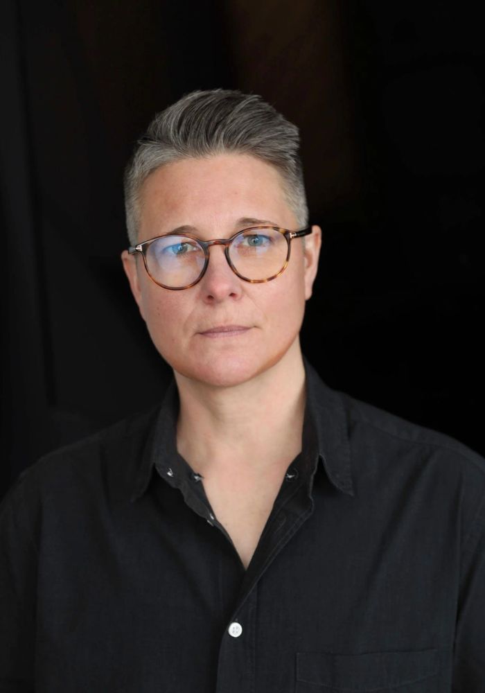 Person with short gray hair and glasses wearing a black shirt against a dark background.