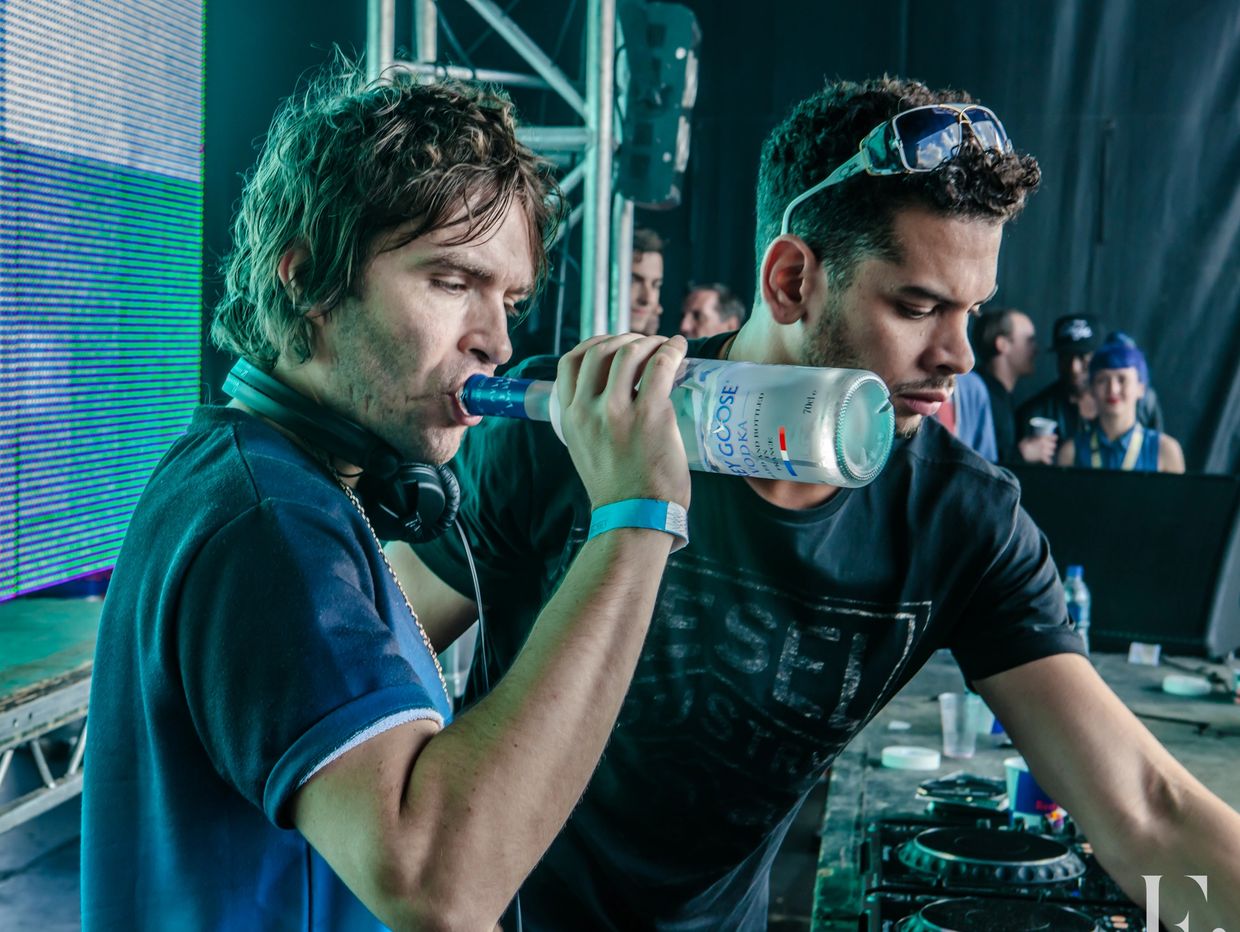 Two men at a DJ booth, one drinking from a vodka bottle.