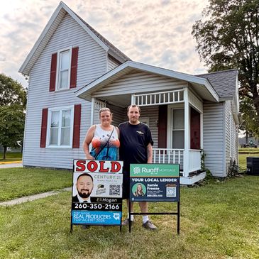Couple standing in front of a sold house with real estate signs.